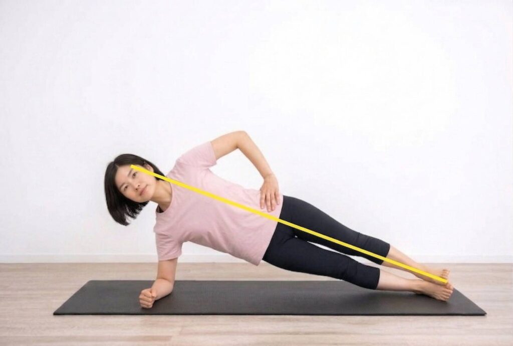 短い黒髪の女性が、明るい部屋の黒いヨガマットの上で、正しいフォームのサイドプランクを行っている。彼女はピンクのTシャツと黒いレギンスを着用し、体は頭から足首まで一直線で、足は積み重ねられている。画像には、彼女の体の上に、正しいアライメントを示す明るい黄色の直線が重ねられている。