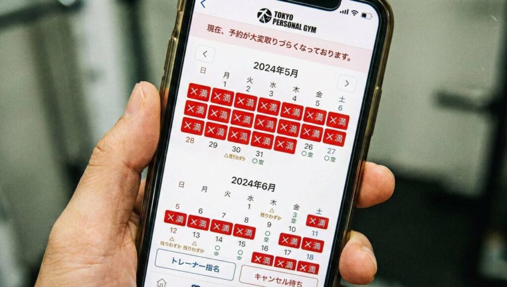 スマートフォンの画面に表示された「TOKYO PERSONAL GYM」の予約システム。2024年5月と6月のカレンダーのほぼ全ての日付に赤色の「× 満」マークが付いており、予約が非常に取りづらい満席状態であることが示されている。画面上部には「現在、予約が大変取りづらくなっております。」というメッセージがある。