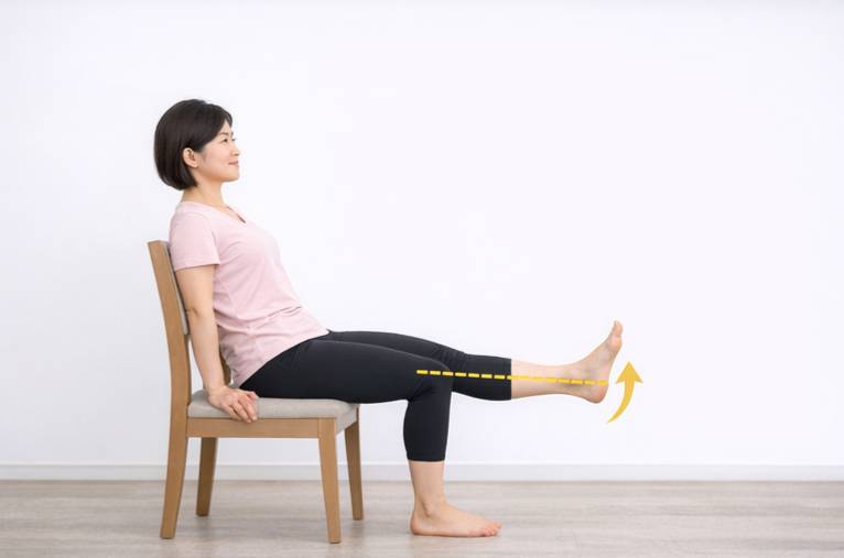 女性が右脚のレッグエクステンションエクササイズを行っている。彼女はピンクのTシャツと黒いクロップドレギンスを着用し、まっすぐ伸ばした脚は床と平行で、足首が曲がっている。彼女の大腿四頭筋(太ももの前部)の上には黄色い点線があり、持ち上げられた足の下には黄色いカーブした矢印があり、大腿四頭筋の強化と足の動きの焦点を強調している。