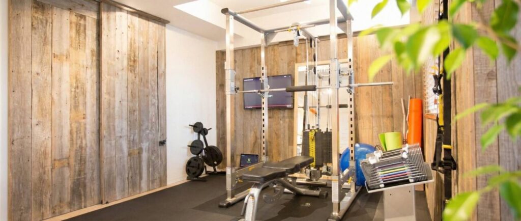 木目調の壁と緑が落ち着いた雰囲気を醸し出す、ビーコンセプトの完全個室トレーニングルーム。パワーラック、トレーニングベンチ、ヨガマットなどの設備が整い、大型の鏡やテレビ、空気清浄機も設置された清潔感のある空間。
